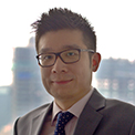 Person wearing glasses and a suit, standing indoors with cityscape in the background.