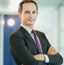 Person in a suit with arms crossed, smiling in an office setting.