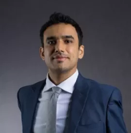Person in a blue suit and tie smiling with a neutral background.