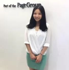 Person smiling in front of PageGroup sign, wearing a white top and green skirt.