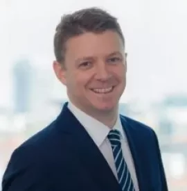 Person in a suit smiling in an office setting with a blurred cityscape in the background.