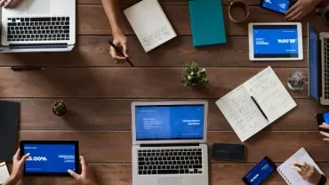 Group of people working on laptops and tablets at a wooden table.