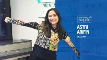 Person smiling with arms outstretched, standing in a lab with shelves and bottles.