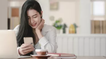 Person smiling at phone in a cozy room with a laptop and coffee on table.