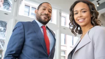 Two people smiling in an office setting with large windows.