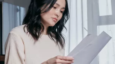 Person seated, holding and reading papers by a window.