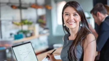 Smiling person with long hair holding a pen in front of a laptop showing graphs in a cafe setting.
