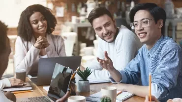 Group of people smiling and talking around a table with laptops and notebooks.