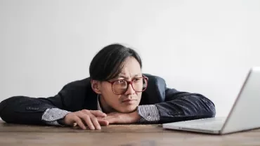Person in a suit resting head on hands, staring at a laptop on a wooden table.