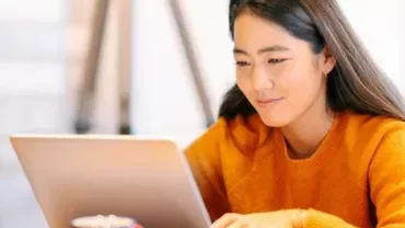 Person in orange sweater working on a laptop at home.