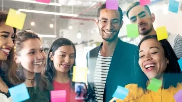 Group of people smiling and looking at sticky notes on a glass wall.