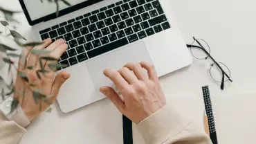 Person working on a laptop at a desk with notebooks and glasses.
