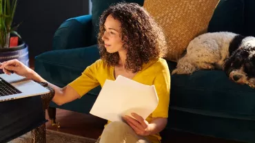 Person working at home on a laptop with a dog resting on a sofa.