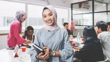 Person smiling in an office with colleagues gathered around a table.