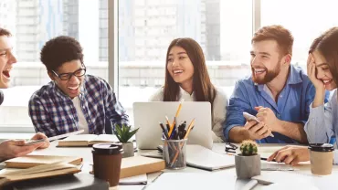 A group of office workers of different ethnicities and in casual wear, chatting and laughing on the same table