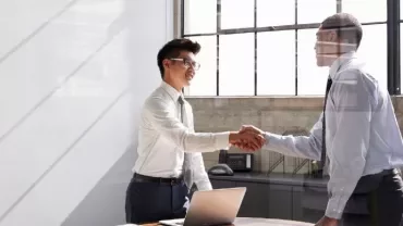 Two people in an office shaking hands by a desk with a laptop.