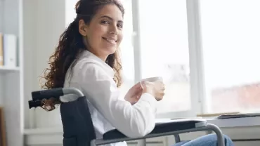 Person in a wheelchair smiling and holding a mug by a window.