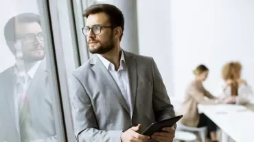 Person in a suit looking out the window, holding a tablet.