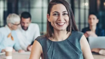 Smiling person in a grey outfit with a group in a meeting room.