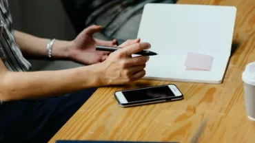 Person holding a pen over an open notebook with a pink sticky note.