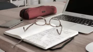 Eyeglasses resting on an open notebook with a laptop and pencil case on a wooden desk.