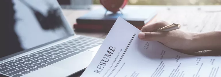 Person holding a resume next to a laptop on a wooden desk.
