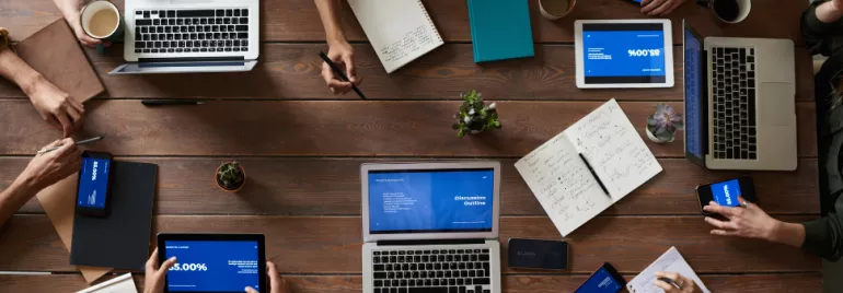 Group of people working on laptops and tablets at a wooden table.