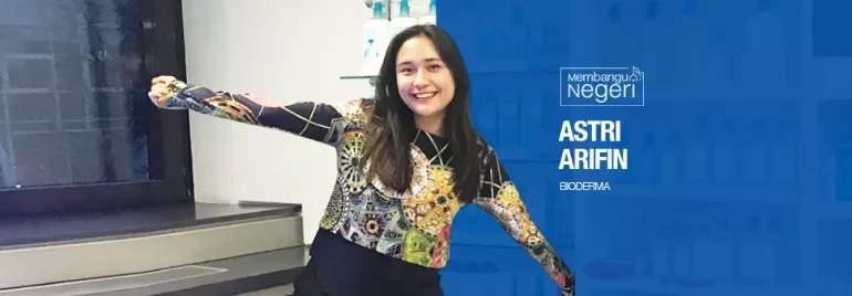 Person smiling with arms outstretched, standing in a lab with shelves and bottles.