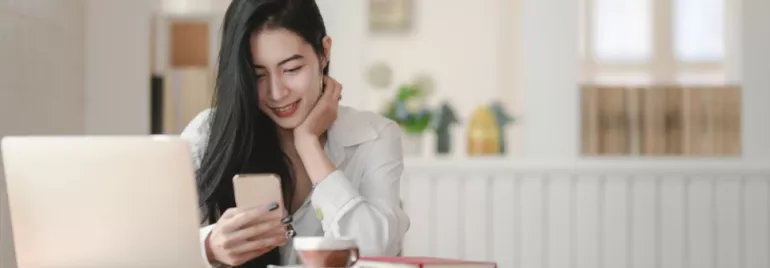 Person smiling at phone in a cozy room with a laptop and coffee on table.