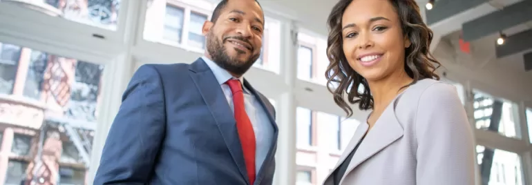 Two people smiling in an office setting with large windows.