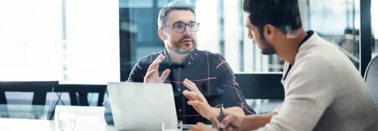 Two people have a discussion in an office with a laptop and notepad.
