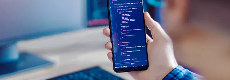 Person holding a phone displaying code, with a blurred computer screen in the background.