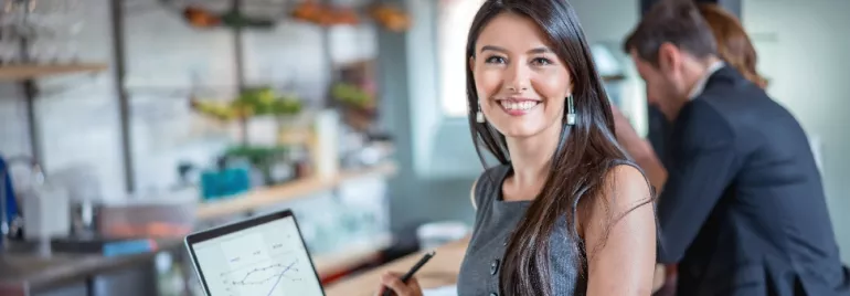 Smiling person with long hair holding a pen in front of a laptop showing graphs in a cafe setting.