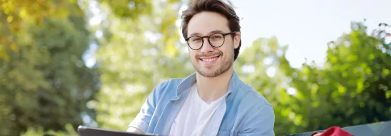 One Caucasian man in casual clothes holding a laptop and sitting on a garden bench and smiling to camera