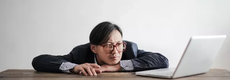 Person in a suit resting head on hands, staring at a laptop on a wooden table.