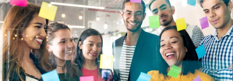 Group of people smiling and looking at sticky notes on a glass wall.