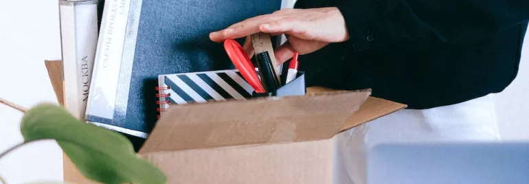Person packing books and office supplies into a cardboard box.