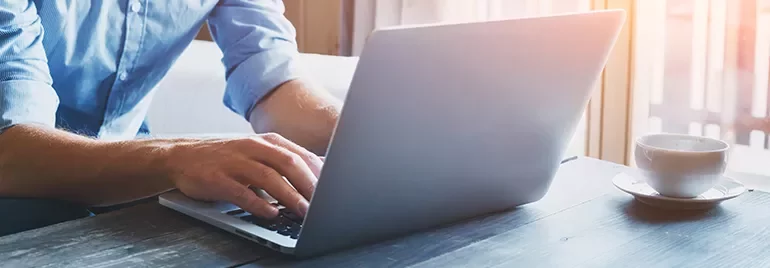 Person typing on a laptop at a wooden table with a cup of coffee beside them.