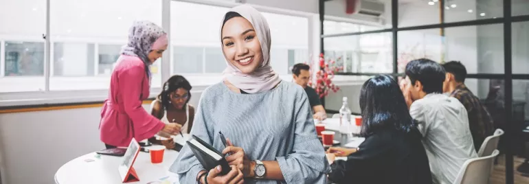 Person smiling in an office with colleagues gathered around a table.