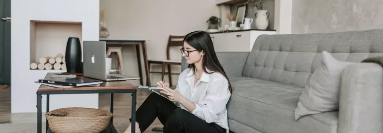 Person sitting on floor in living room, working on a laptop and taking notes.