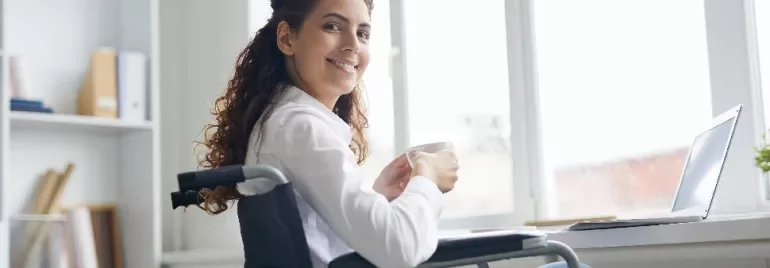 Person in a wheelchair smiling and holding a mug by a window.