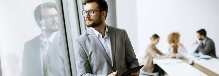 Person in a suit looking out the window, holding a tablet.
