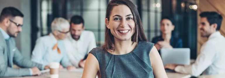 Smiling person in a grey outfit with a group in a meeting room.