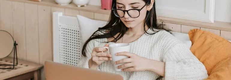 Person sitting on a cozy couch, holding a coffee mug while using a laptop.