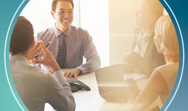 A group of people in business attire having a meeting around a table, with sunlight streaming in.