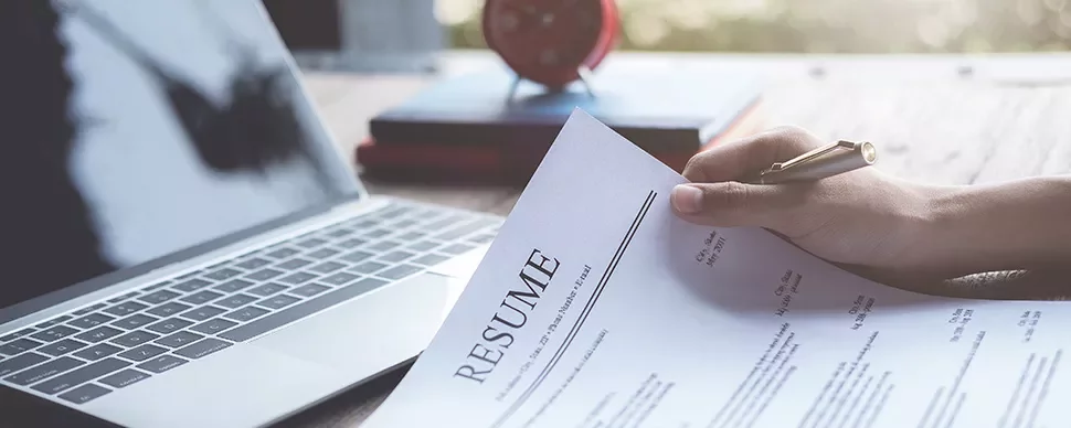 Person holding a resume next to a laptop on a wooden desk.