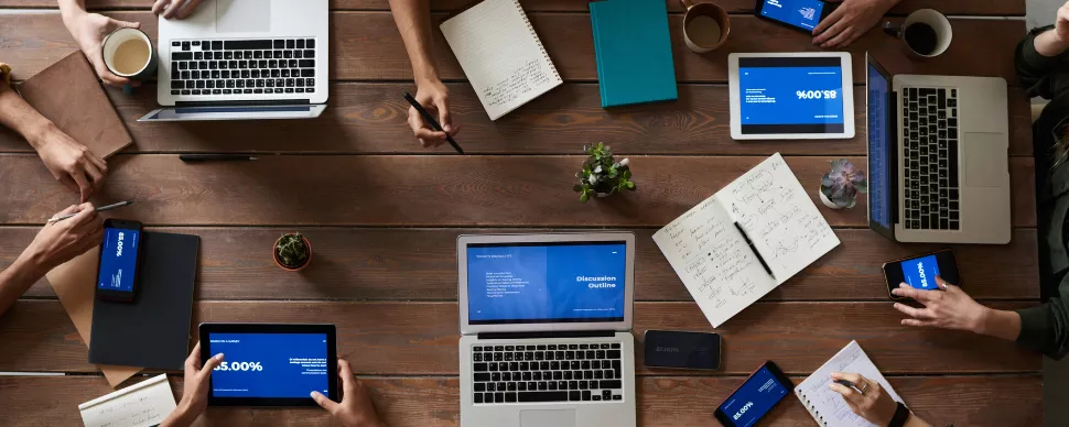 Group of people working on laptops and tablets at a wooden table.