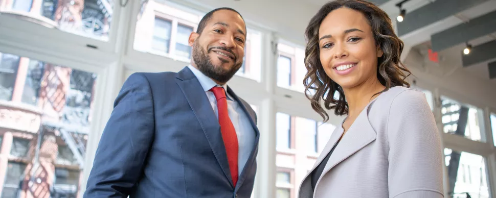 Two people smiling in an office setting with large windows.
