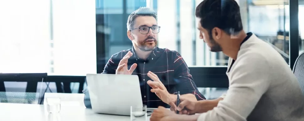 Two people have a discussion in an office with a laptop and notepad.