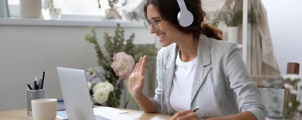 Person wearing headphones, smiling and waving at a laptop screen in a bright room.